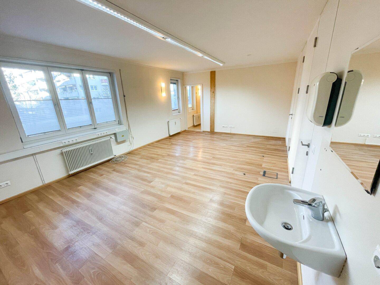 Geräumiger Büroraum mit Laminatboden, großen Fenstern und integriertem Waschbecken und Spinden.