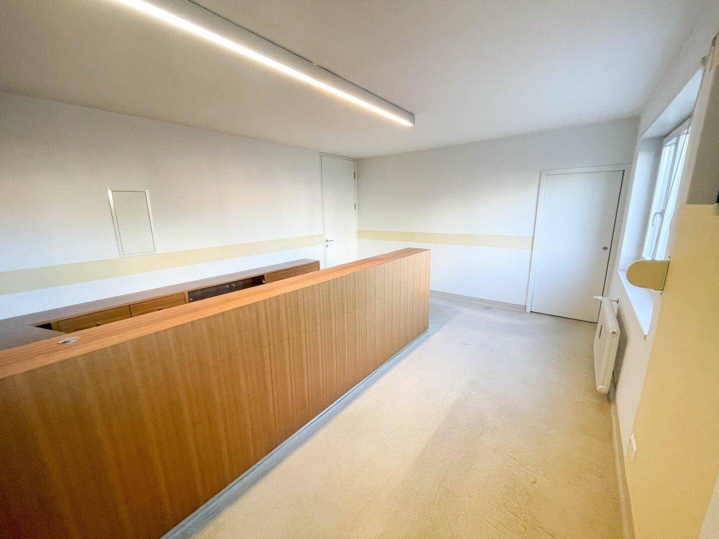 Heller Büroraum mit Holztheke, weißen Wänden und großen Fenstern, ideal für Empfangsbereich.