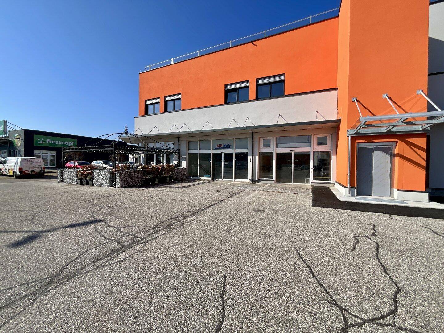 Ansicht des Geschäftsgebäudes mit orangefarbener Fassade, Glasfronten und großem Kundenparkplatz.
