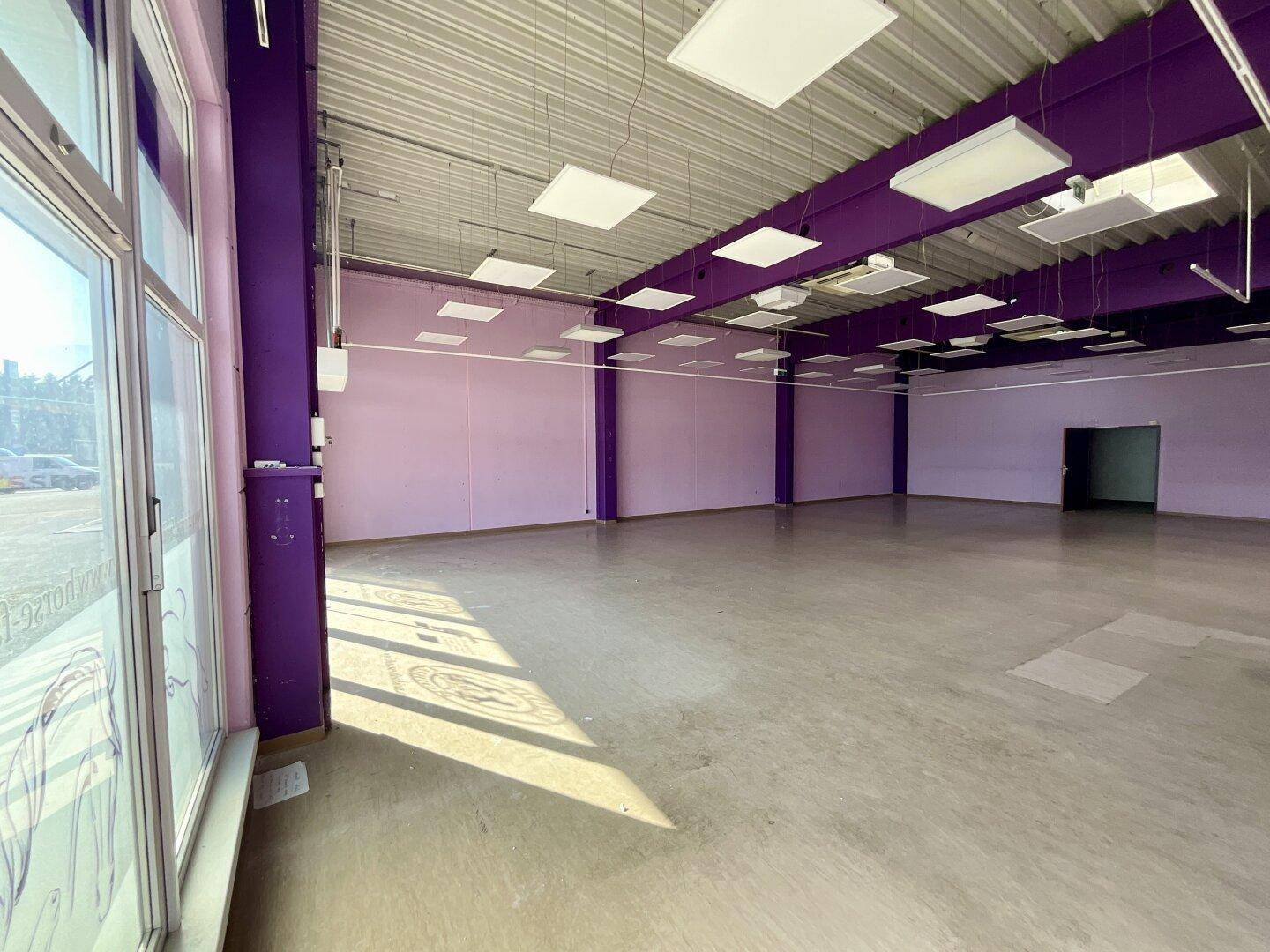 Heller Ladenbereich mit lila Wänden, Betonboden und großen Schaufenstern, die viel Licht hereinlassen.