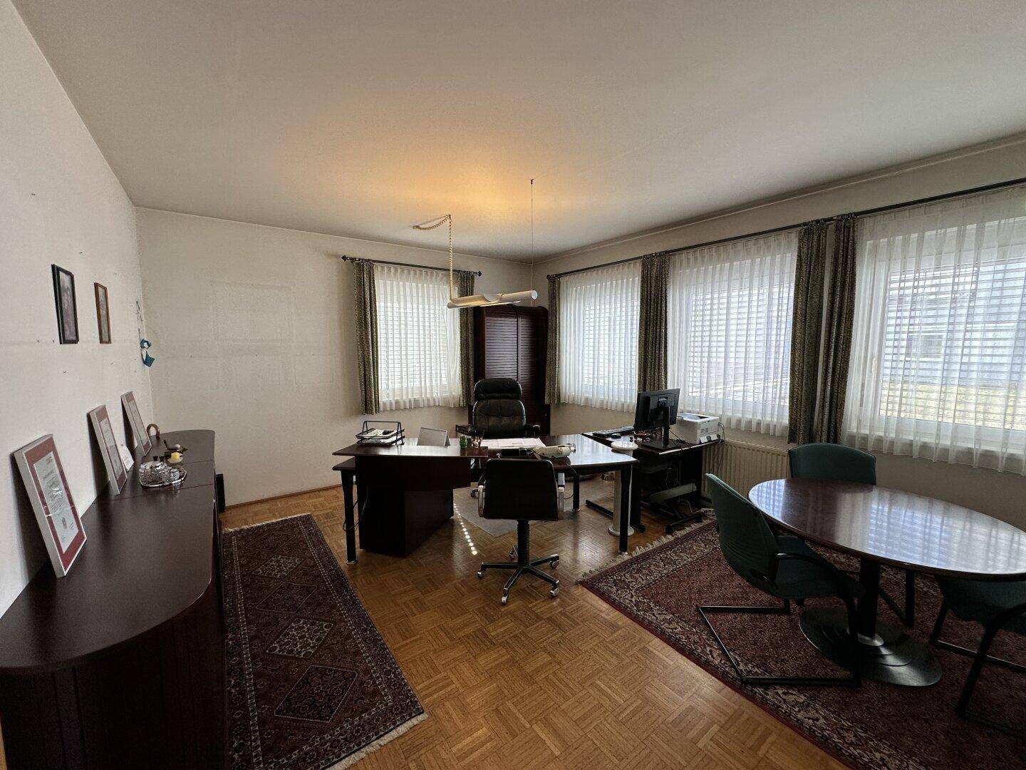 Geräumiges Einzelbüro mit Schreibtisch, Besprechungstisch und großen Fenstern mit Vorhängen.
