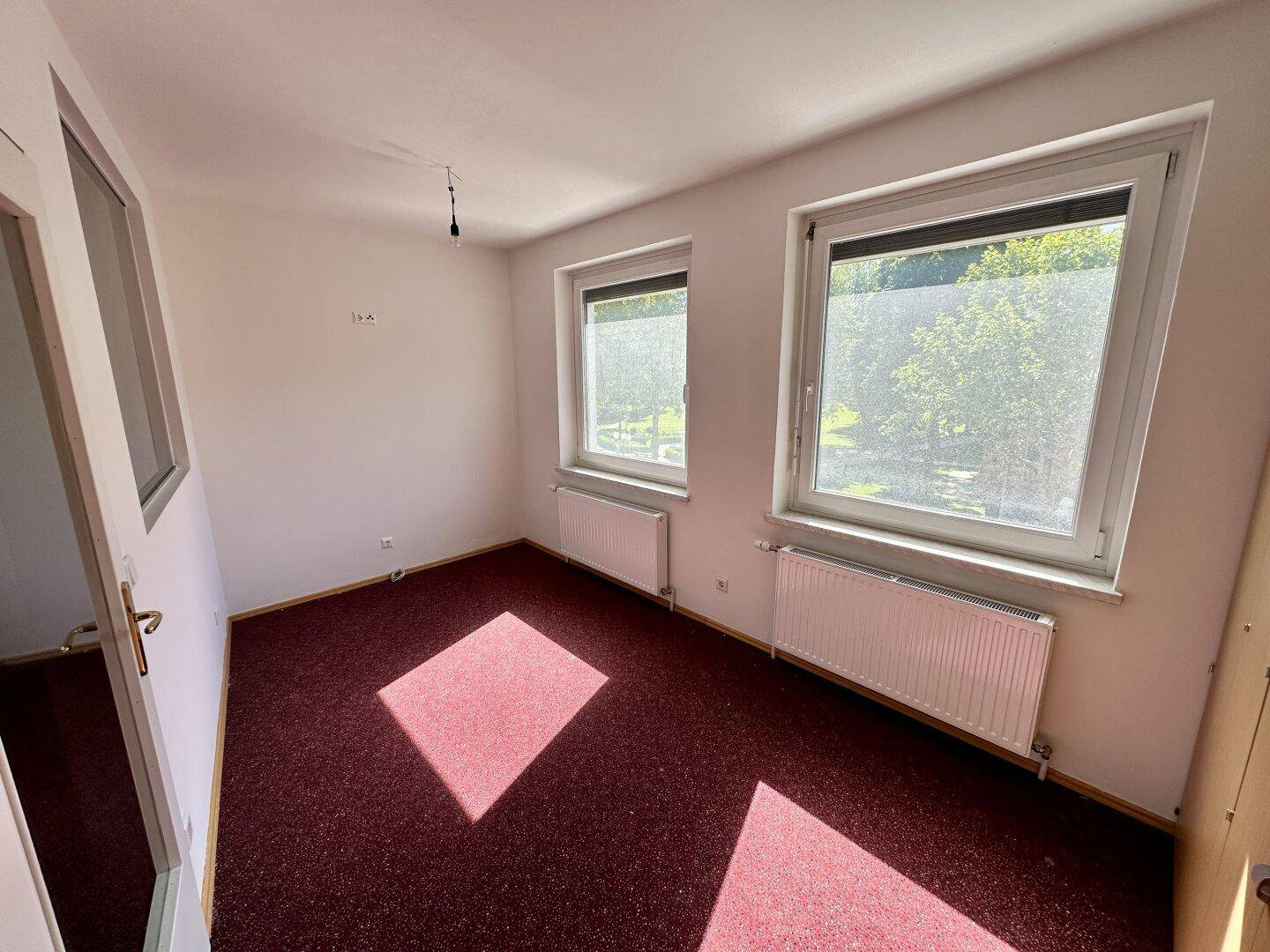 Heller Raum mit rotem Teppichboden und zwei Fenstern, die viel Tageslicht hereinlassen, ideal als Büro oder Schlafzimmer.