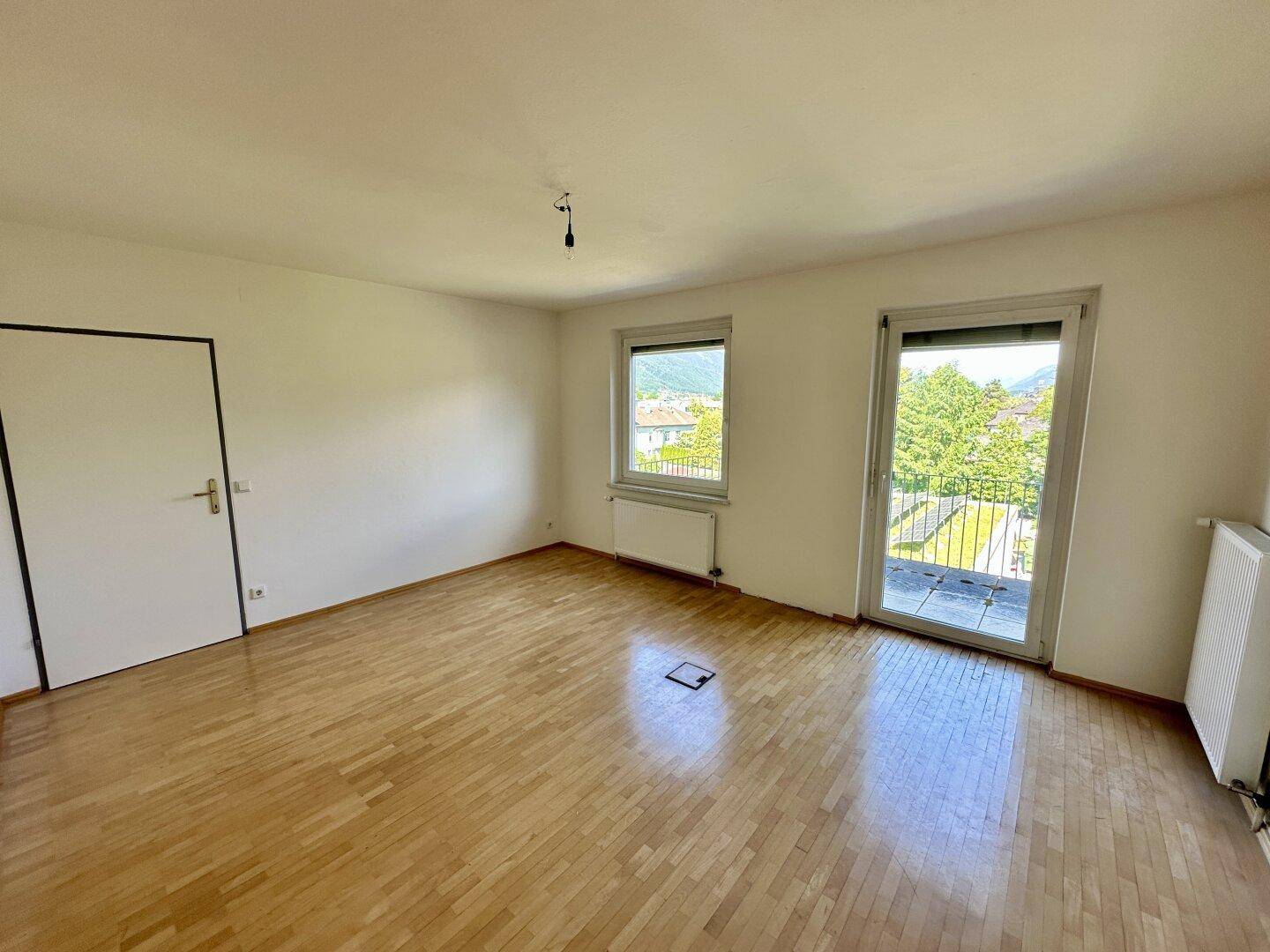 Heller, leerer Raum mit Holzboden, weißen Wänden und Zugang zu einem Balkon, der viel Tageslicht hereinlässt.