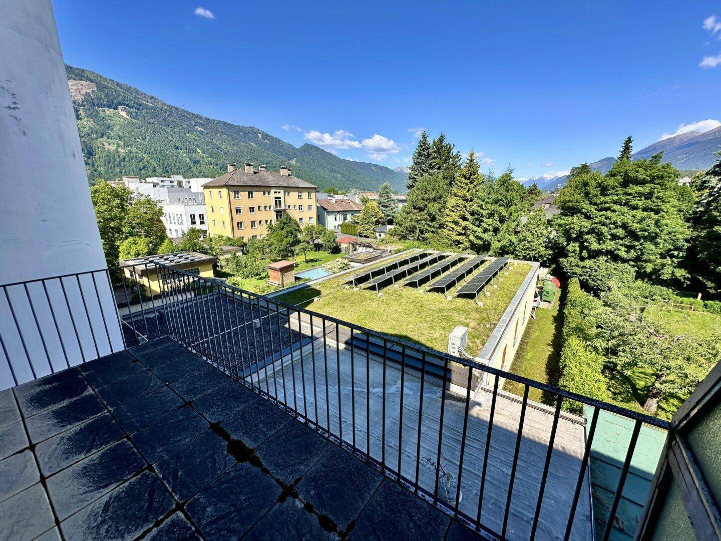 Geräumiger Balkon mit dunklen Fliesen und weitem Blick auf Berge, Bäume und eine begrünte Dachfläche mit Solaranlage.