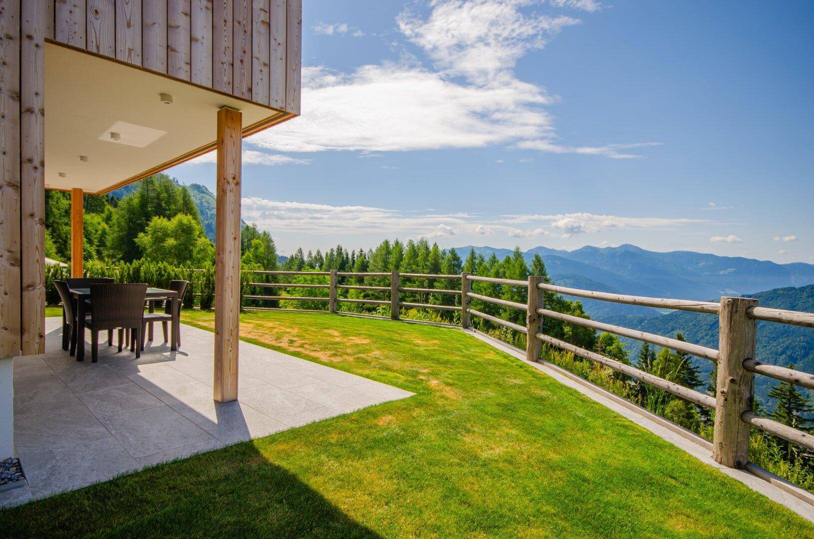 Überdachte Terrasse mit Sitzgelegenheiten und weitläufigem Rasen, umgeben von einer malerischen Berglandschaft.