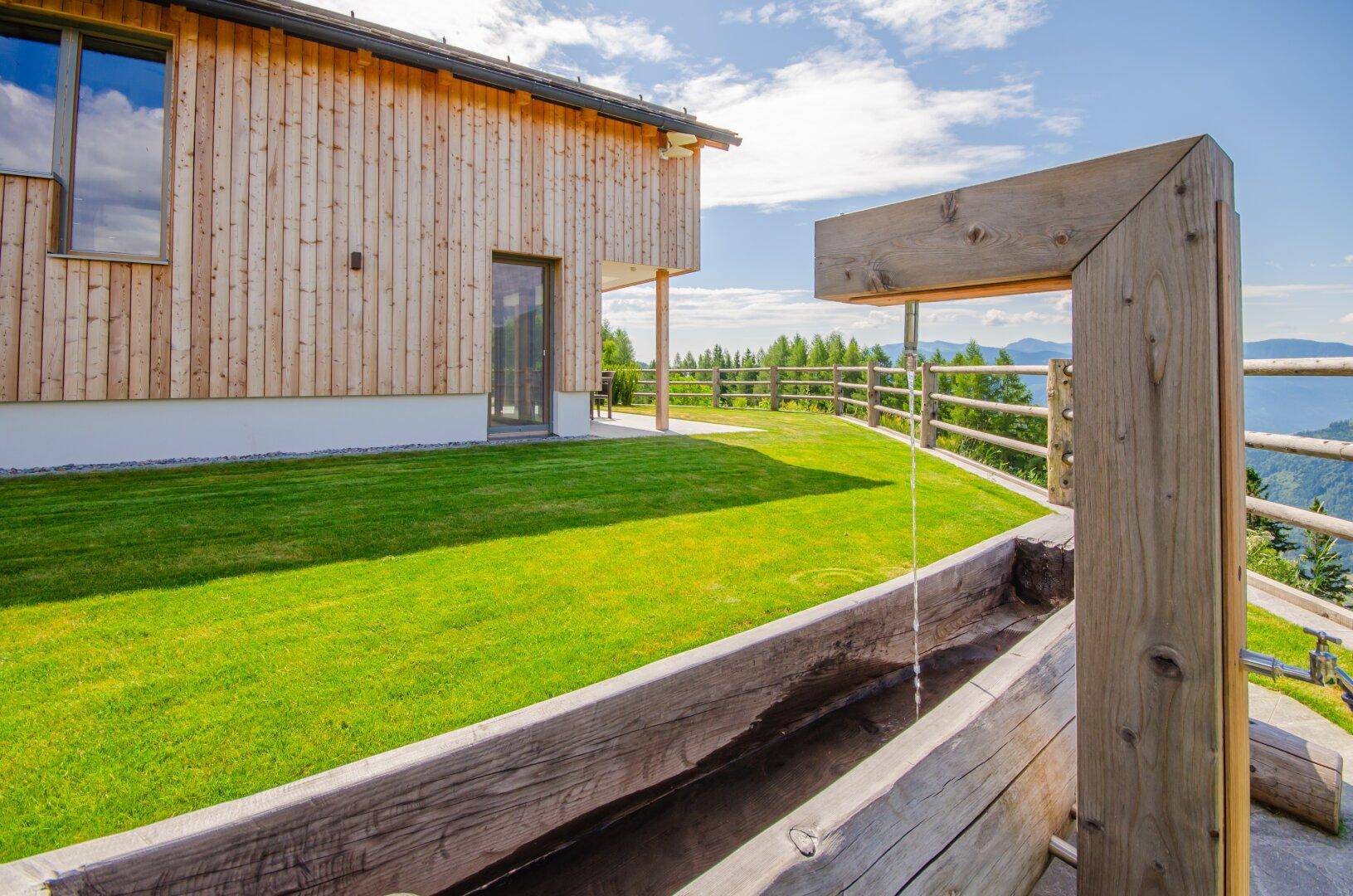 Rustikaler Holzbrunnen im Gartenbereich mit Blick auf die bewaldeten Hügel und das moderne Holzhaus.