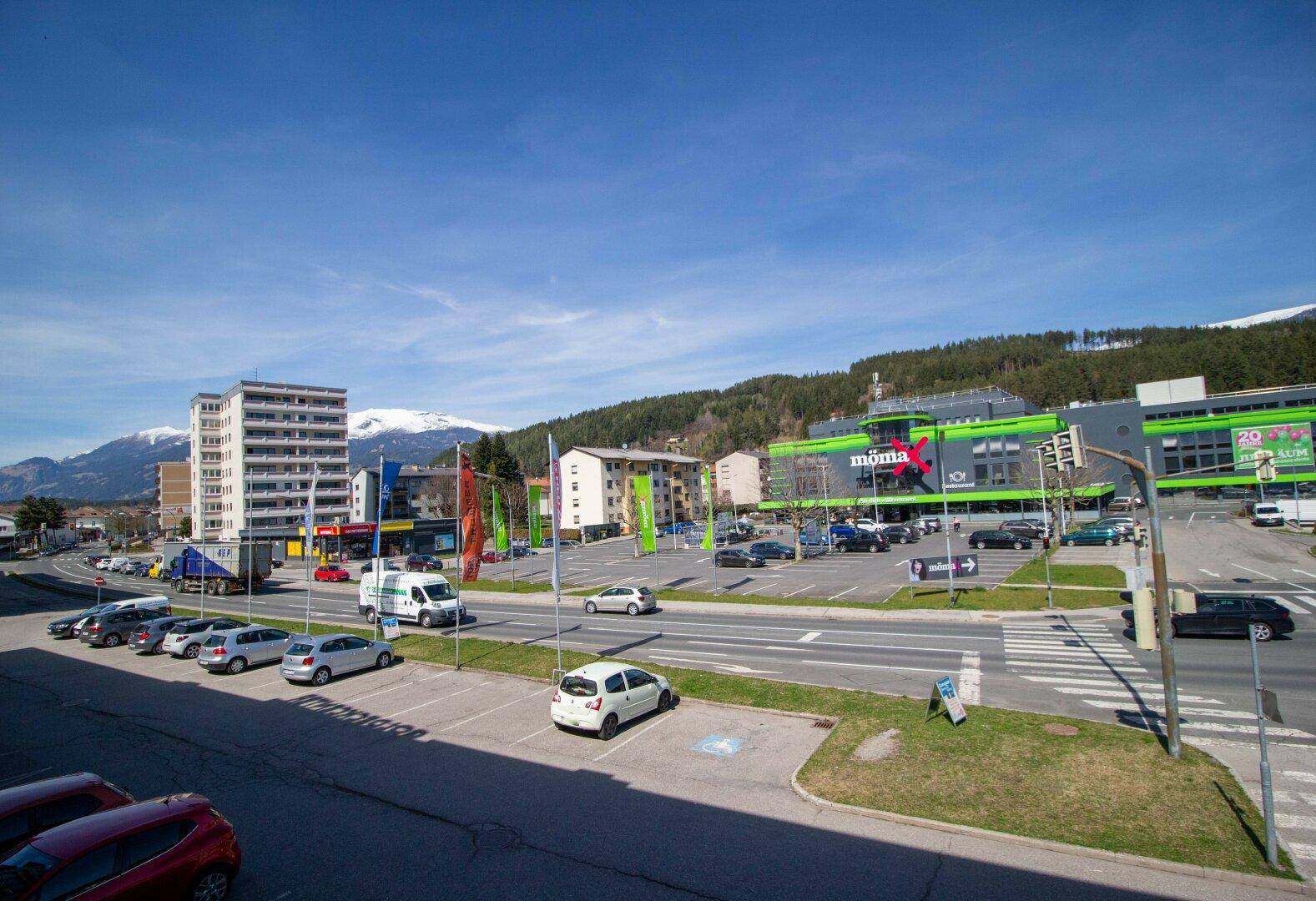 Weitläufige Ansicht der Umgebung mit Parkplätzen, Geschäften und Bergen im Hintergrund unter blauem Himmel.