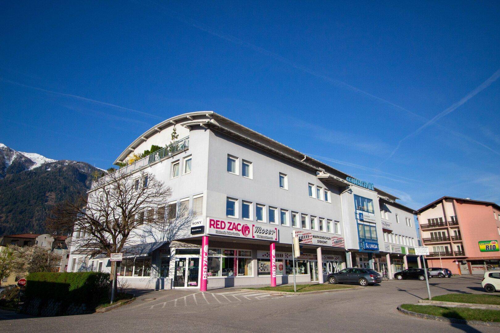 Modernes Geschäftsgebäude mit abgerundeter Fassade und Geschäften im Erdgeschoss, umgeben von Bergen.
