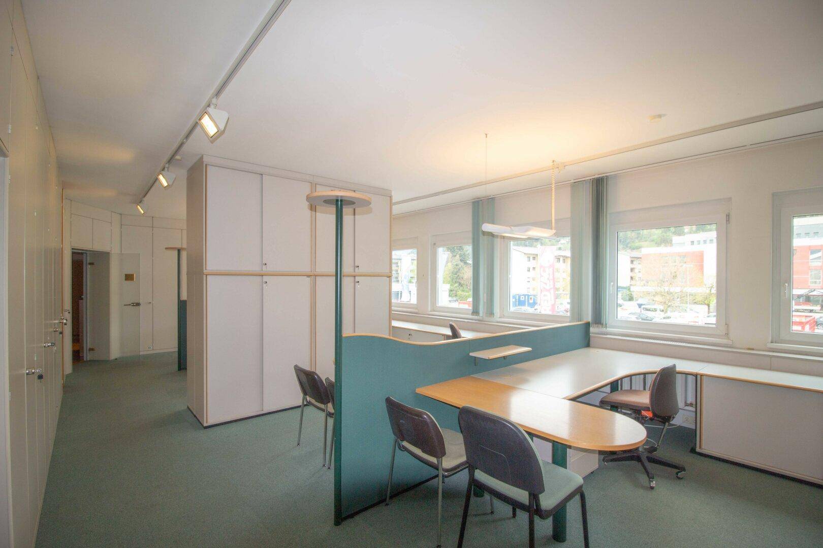 Großraumbüro mit Teppichboden, mehreren Arbeitsplätzen und großen Fenstern, die viel Licht hereinlassen.