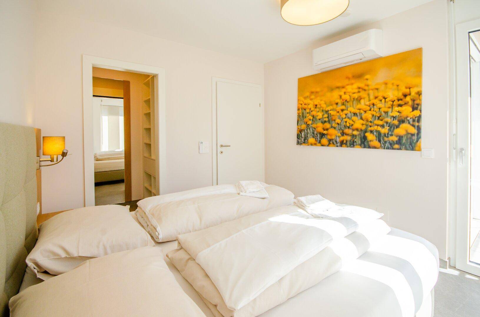 Helles Schlafzimmer mit Doppelbett, Klimaanlage und einem farbenfrohen Wandbild.