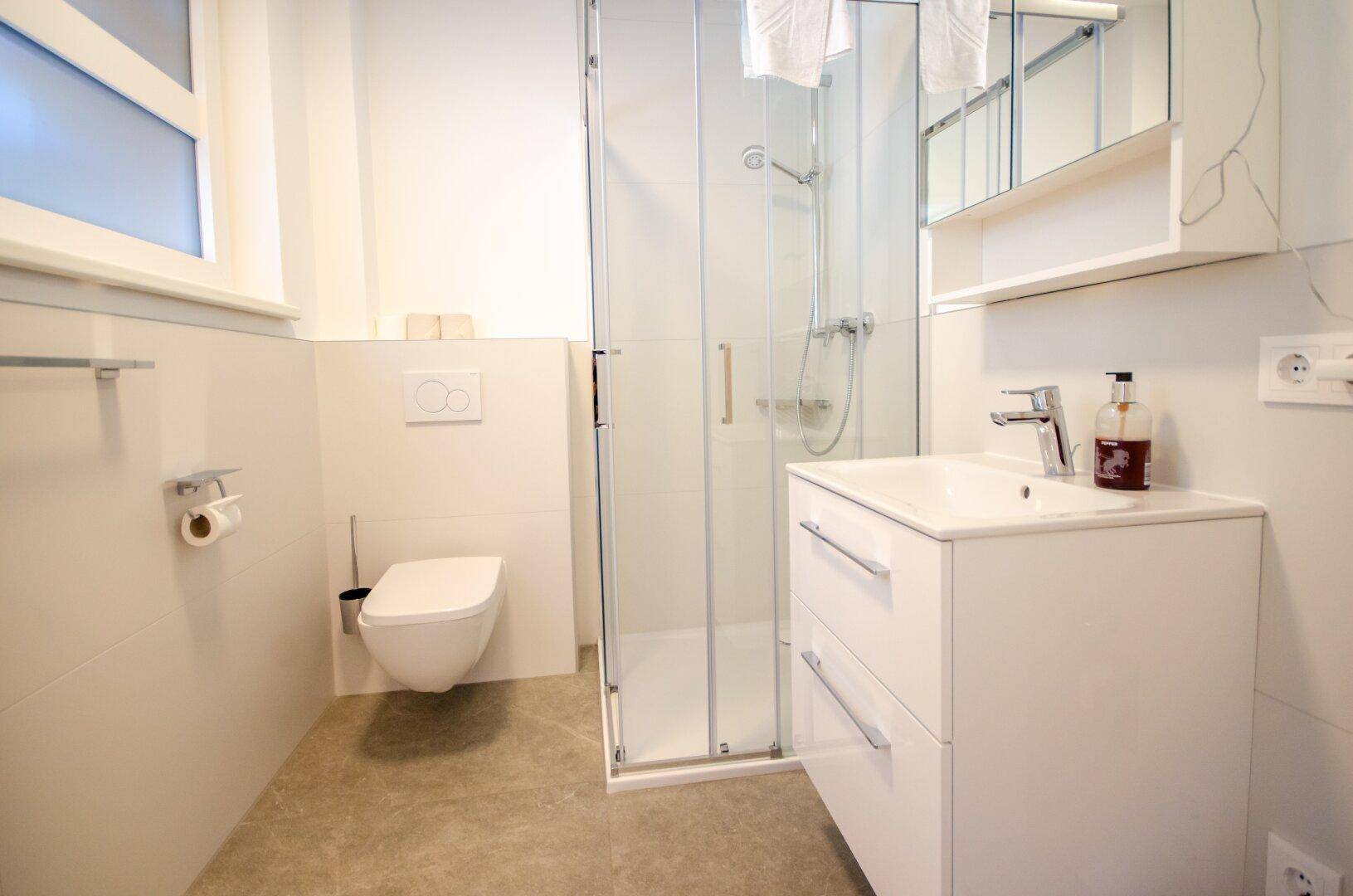 Modernes Badezimmer mit Duschkabine, wandhängendem WC und Fenster für Tageslicht.
