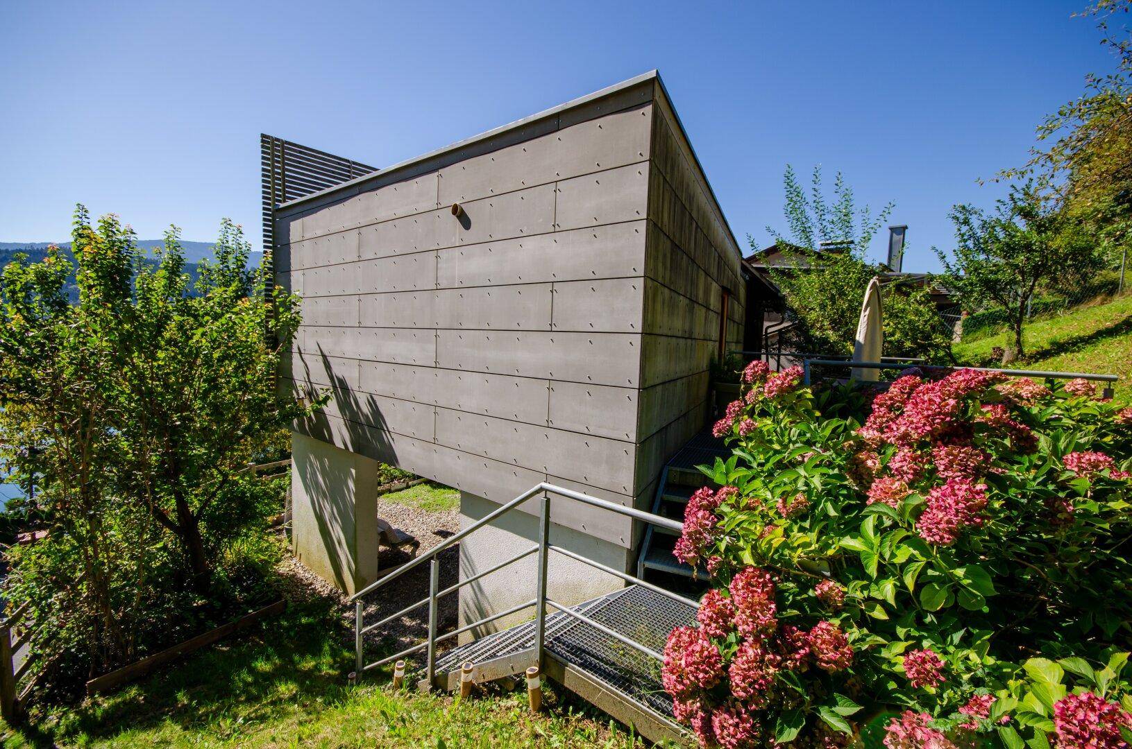 Moderne Architektur mit grauer Fassade und Außentreppe, umgeben von grüner Natur und Blumen.