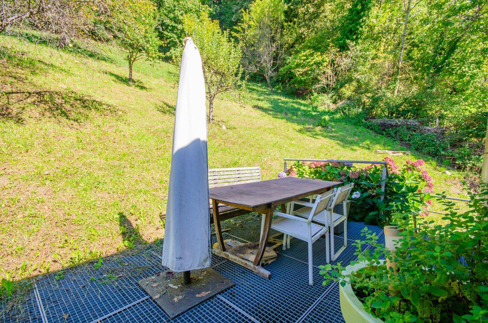 Sonniger Außenbereich mit Esstisch, Stühlen und großem Sonnenschirm im Grünen.