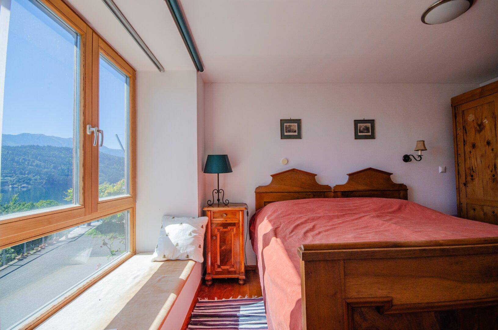 Schlafzimmer mit Holzbett, Fensterbank und herrlichem Blick auf den See und die Berge.