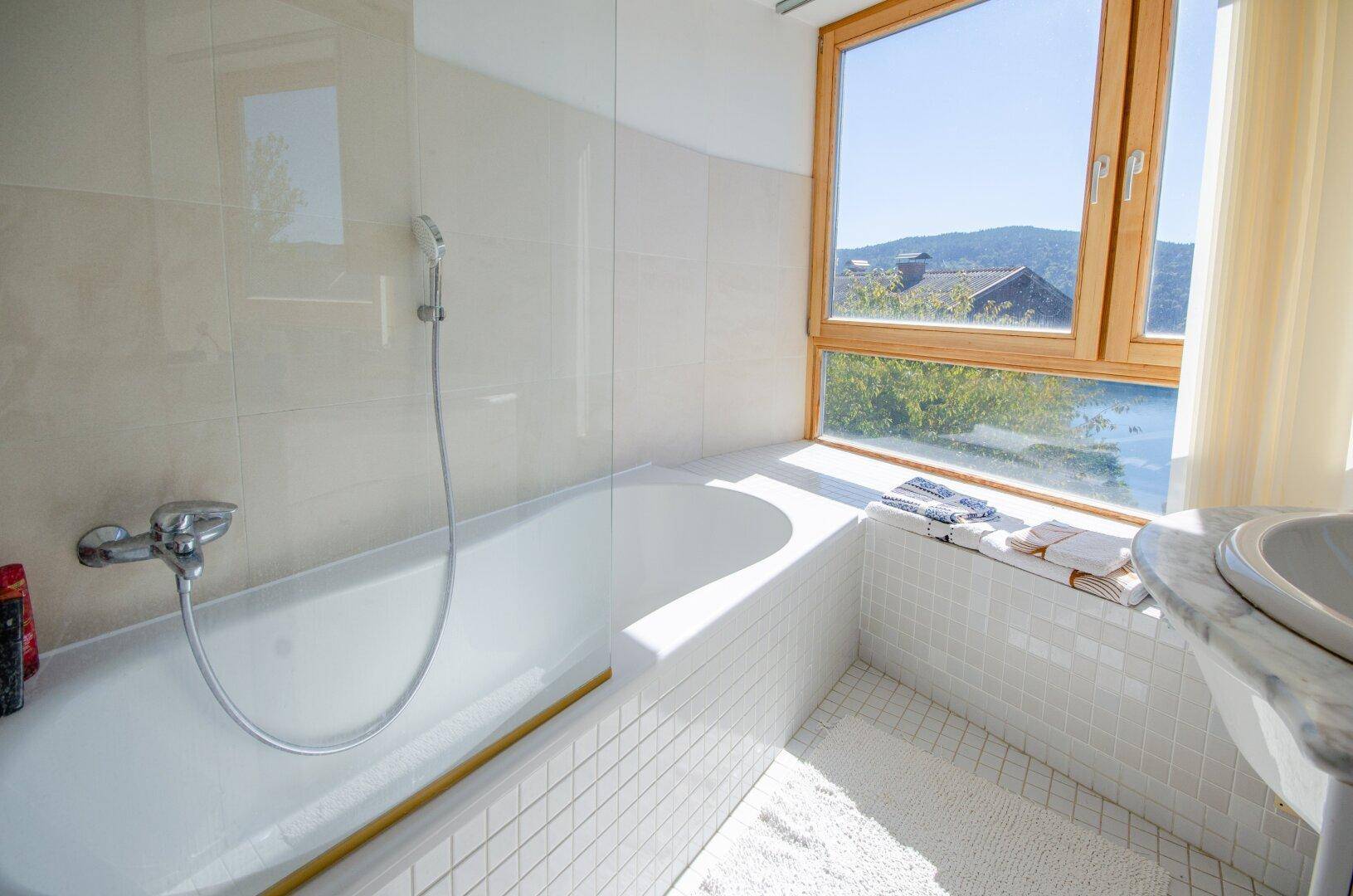 Helles Badezimmer mit Badewanne, weißen Fliesen und Fenster mit Ausblick in die Natur.