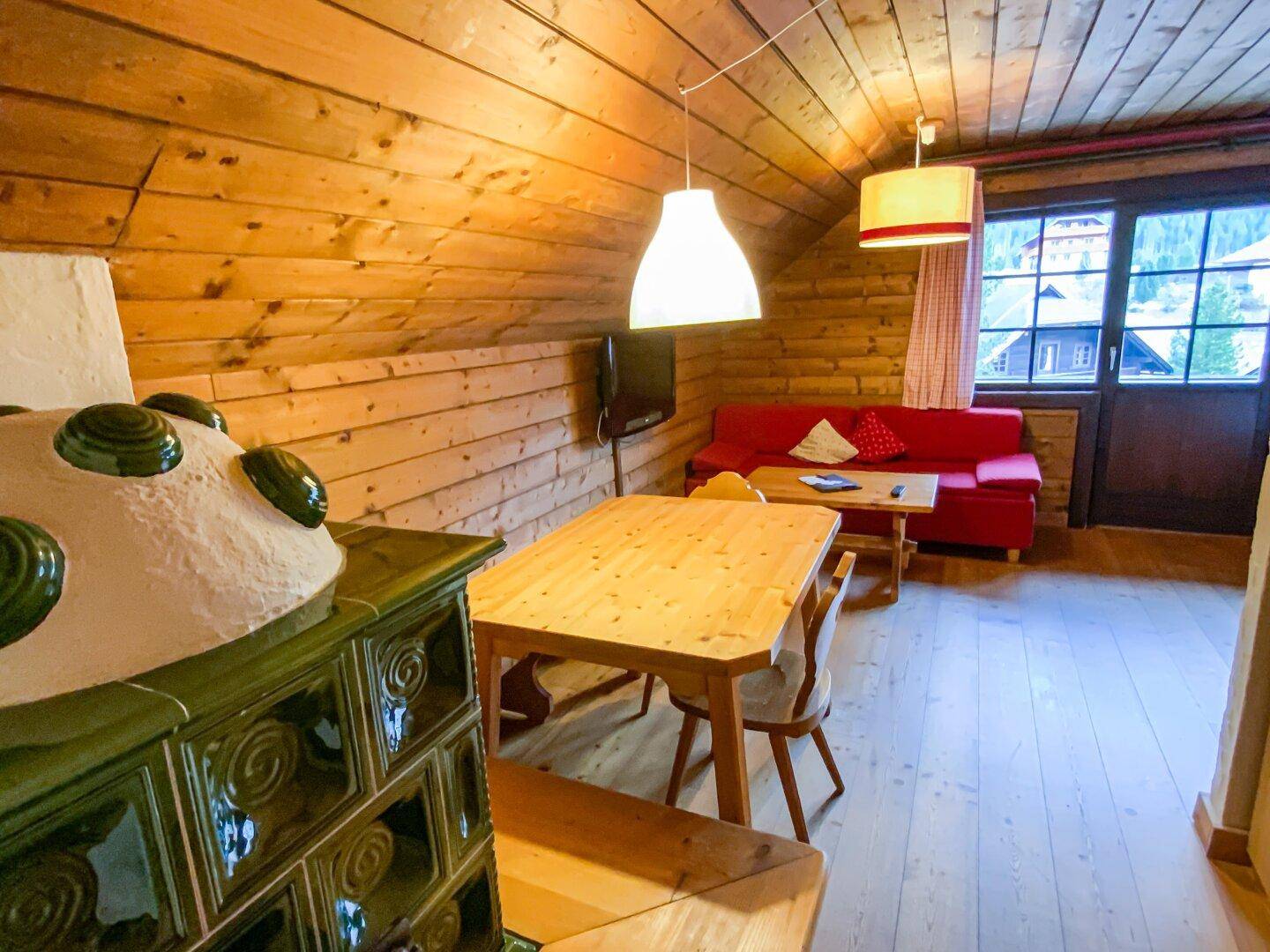 Gemütlicher Wohn-Essbereich mit Holztäfelung, traditionellem Kachelofen und rotem Sofa.