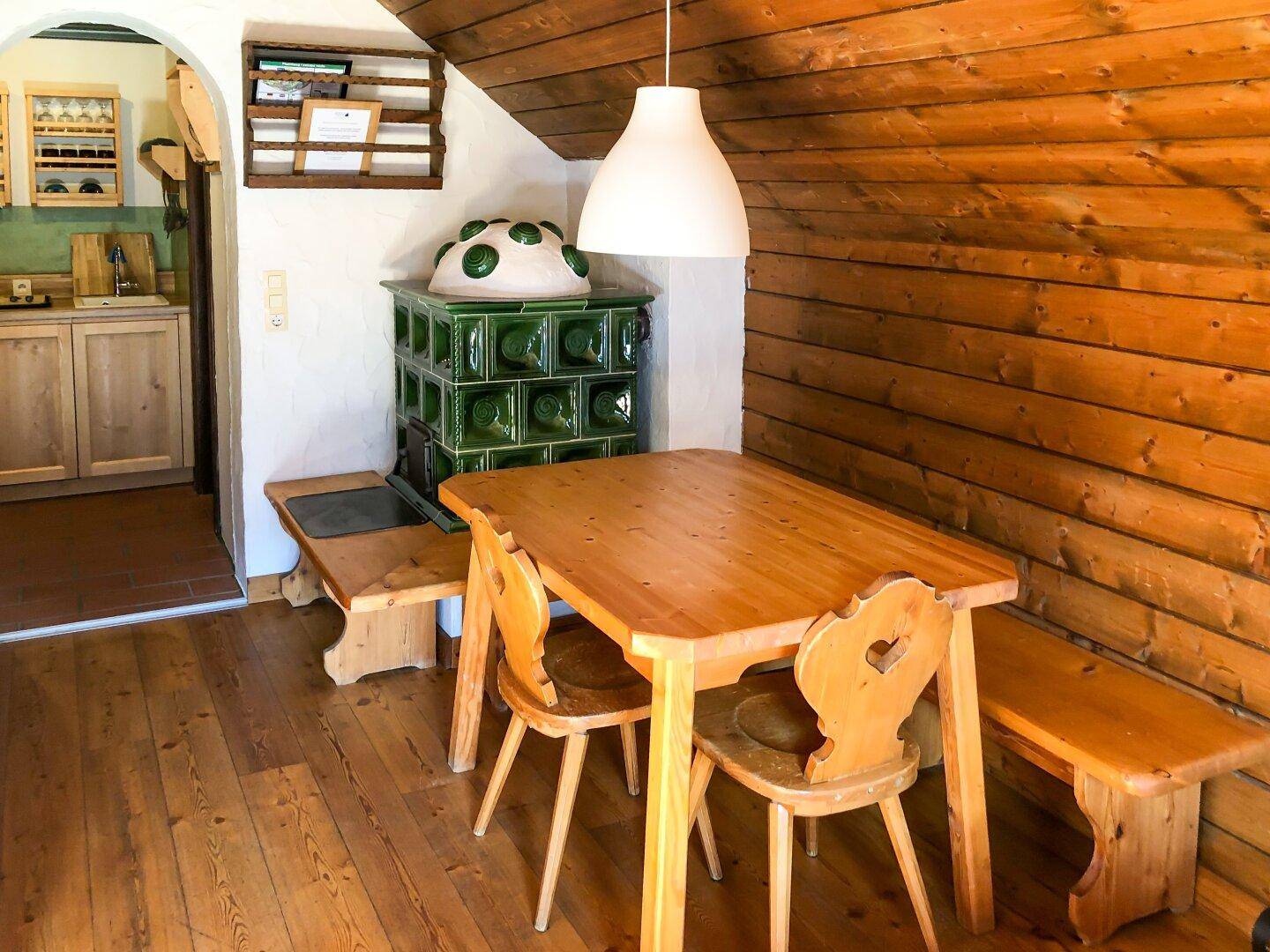Rustikaler Essbereich mit Holztisch, Bänken und einem dekorativen grünen Kachelofen.