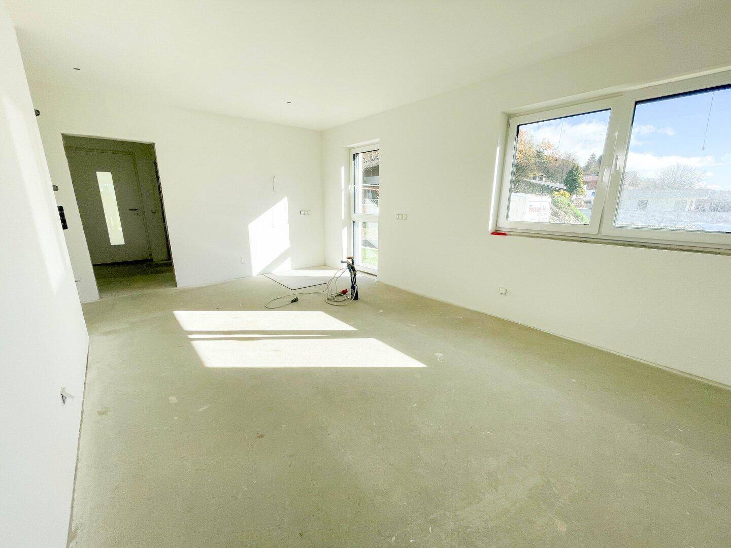 Leerer Raum im Rohbau mit großen Fenstern und Betonboden, bereit für den Ausbau.