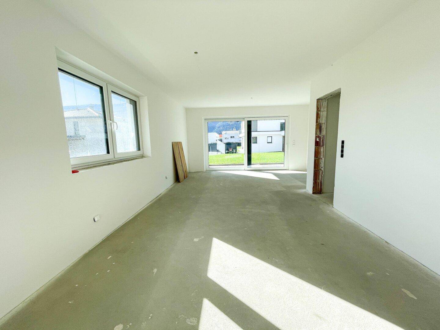 Leerer Raum im Rohbau mit Betonboden, großem Fenster und Schiebetür zum Außenbereich.