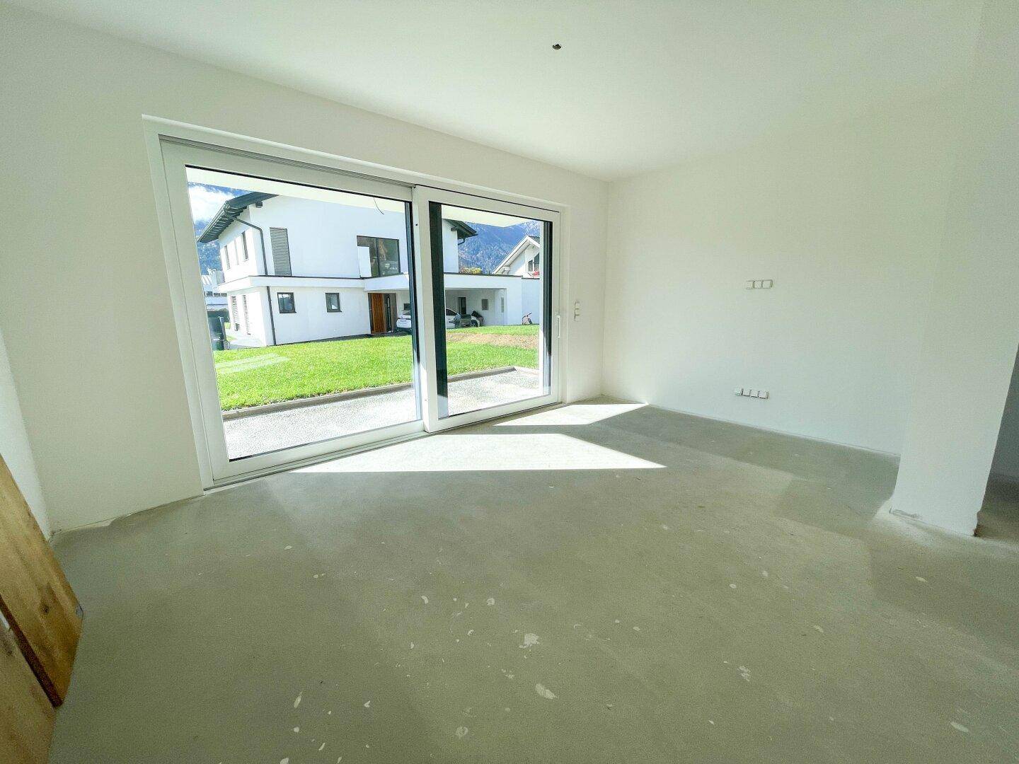 Leerer Raum im Rohbau mit großer Schiebetür zum Garten und Betonboden.