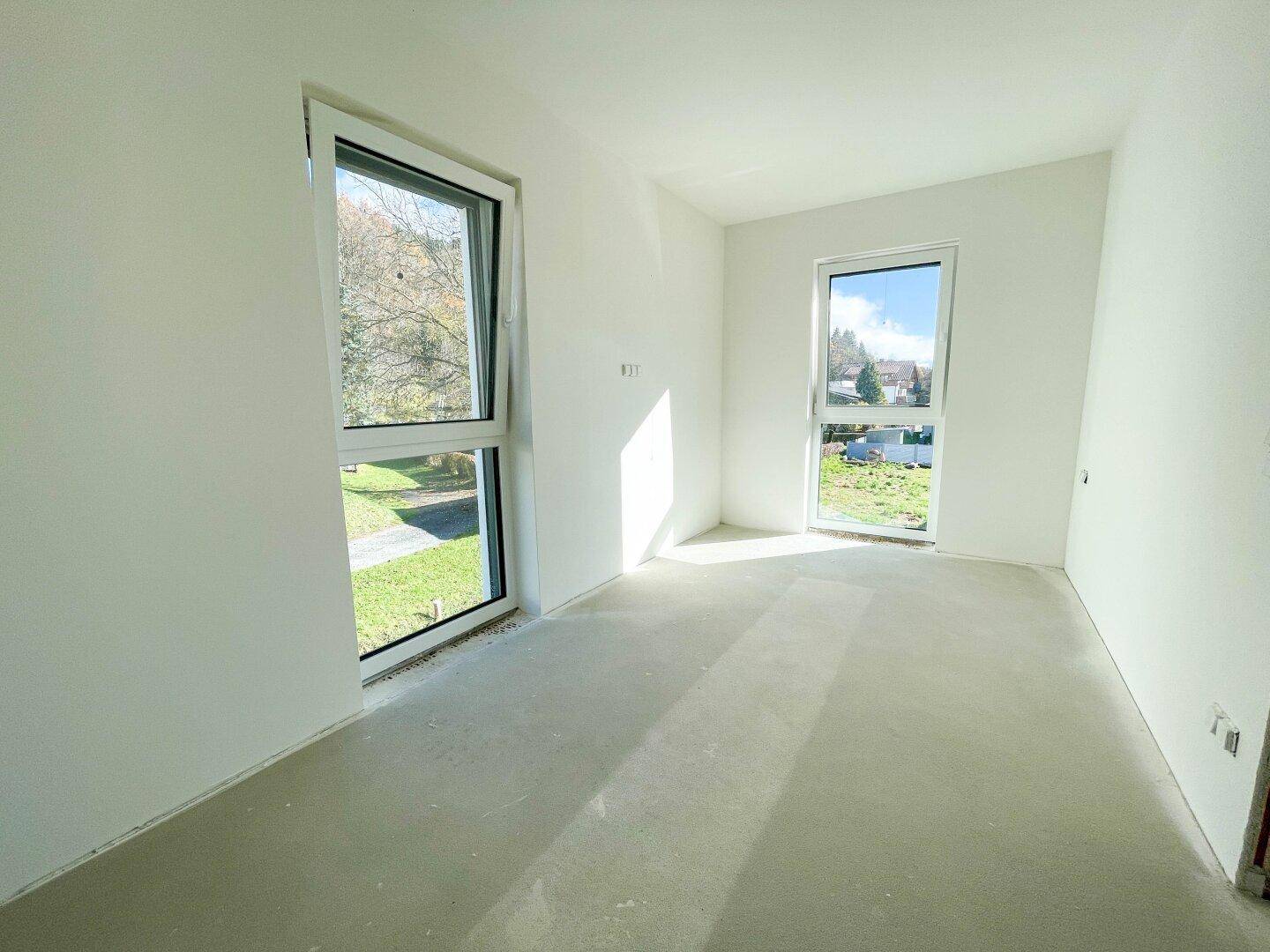 Leerer Raum im Rohbau mit zwei Fenstern und Betonboden, ideal für individuelle Gestaltung.