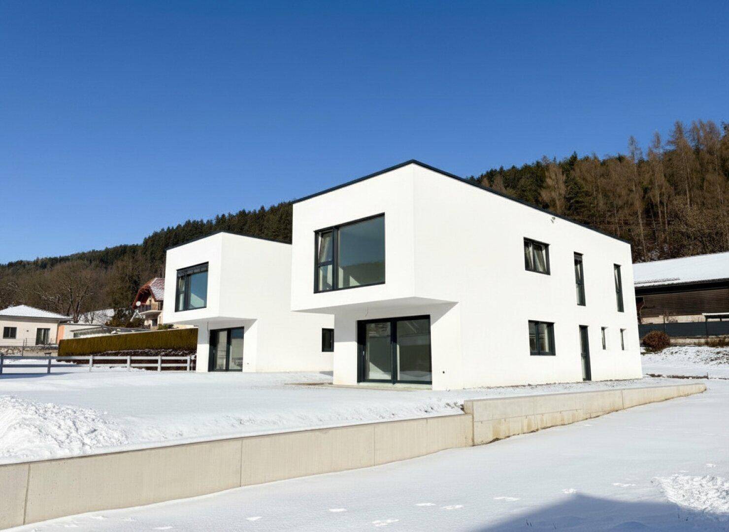 Zwei moderne weiße Gebäude mit großen Fenstern in einer winterlichen Umgebung mit Wald im Hintergrund.