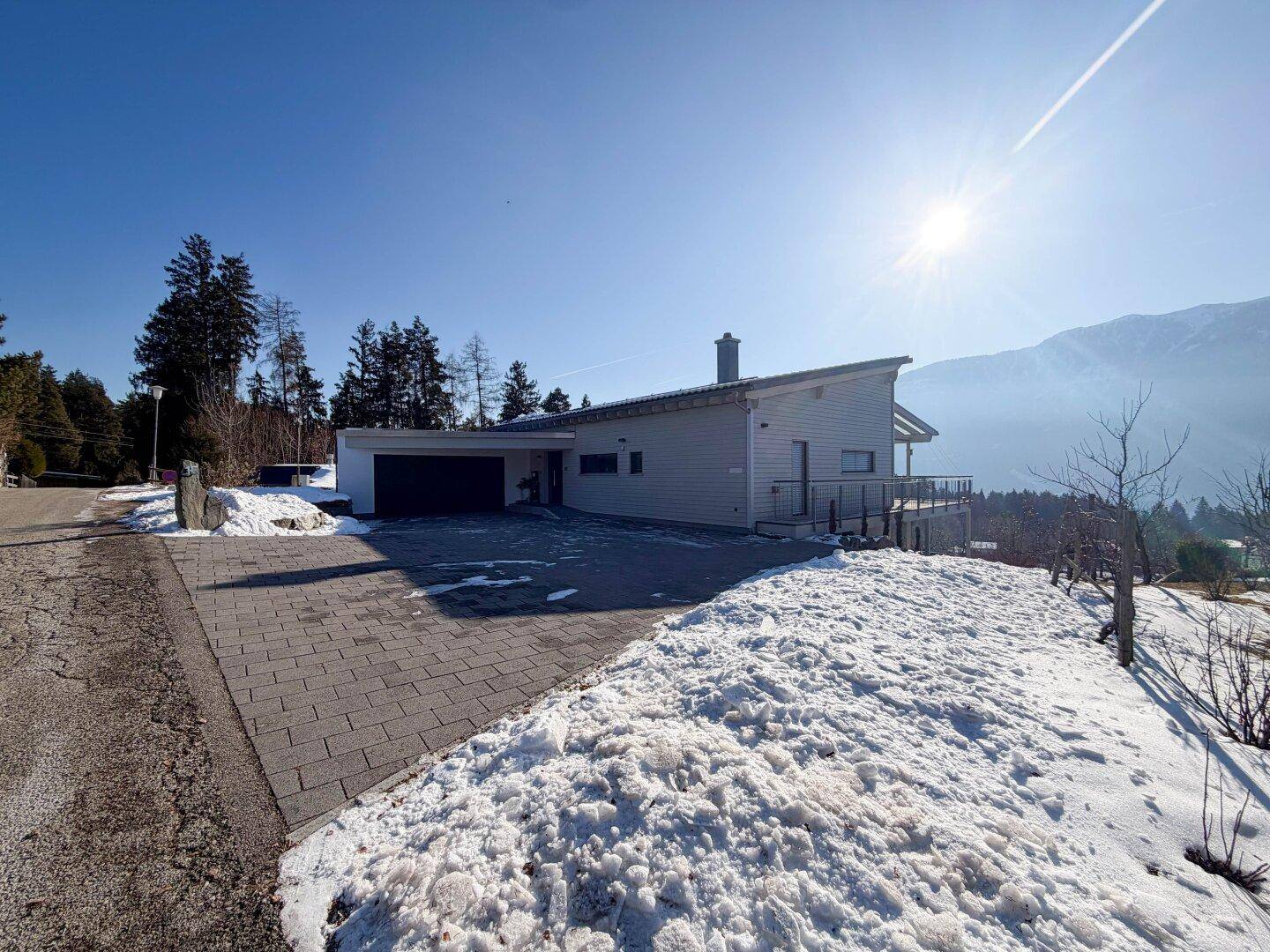 Modernes Haus mit Schrägdach, gepflasterter Auffahrt und schneebedeckter Umgebung unter blauem Himmel.