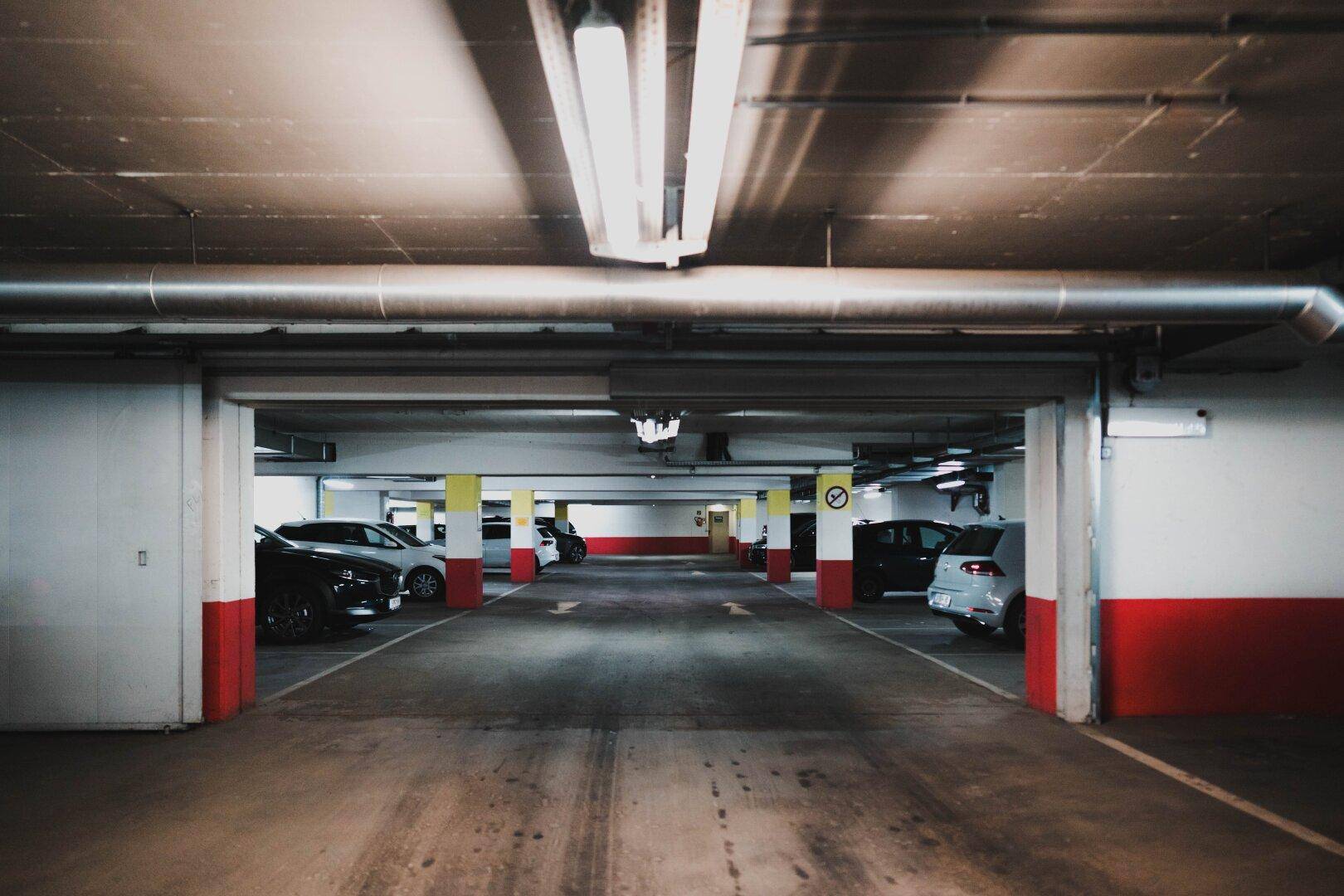 Tiefgarage mit Betonboden, sichtbaren Rohren, Leuchtstoffröhren und geparkten Fahrzeugen.
