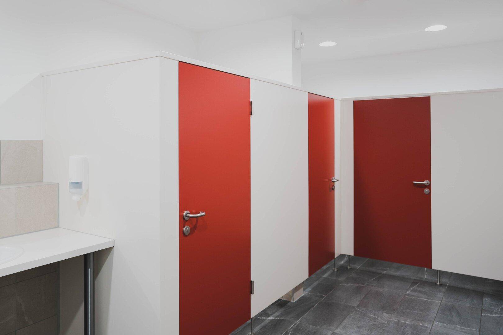 Öffentliche Toilettenanlage mit weißen Trennwänden, roten Türen, Waschbecken und dunklem Fliesenboden.