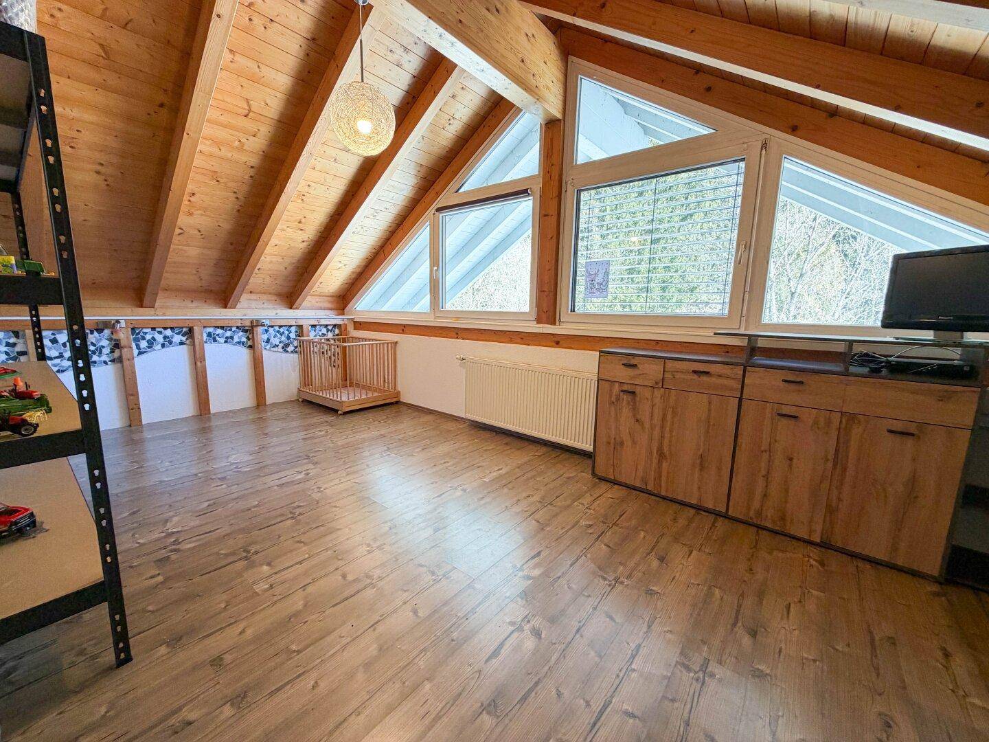 Helles Dachzimmer mit Holzboden, Dachschrägen und großen Fenstern mit Waldblick.