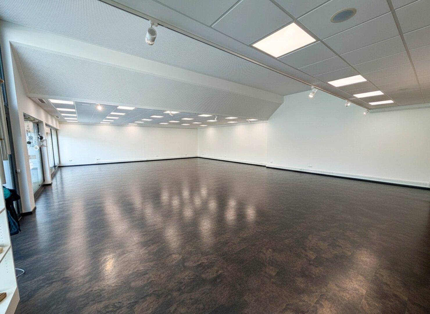 Großer, leerer Gewerberaum mit dunklem Boden, weißen Wänden und moderner Deckenbeleuchtung.