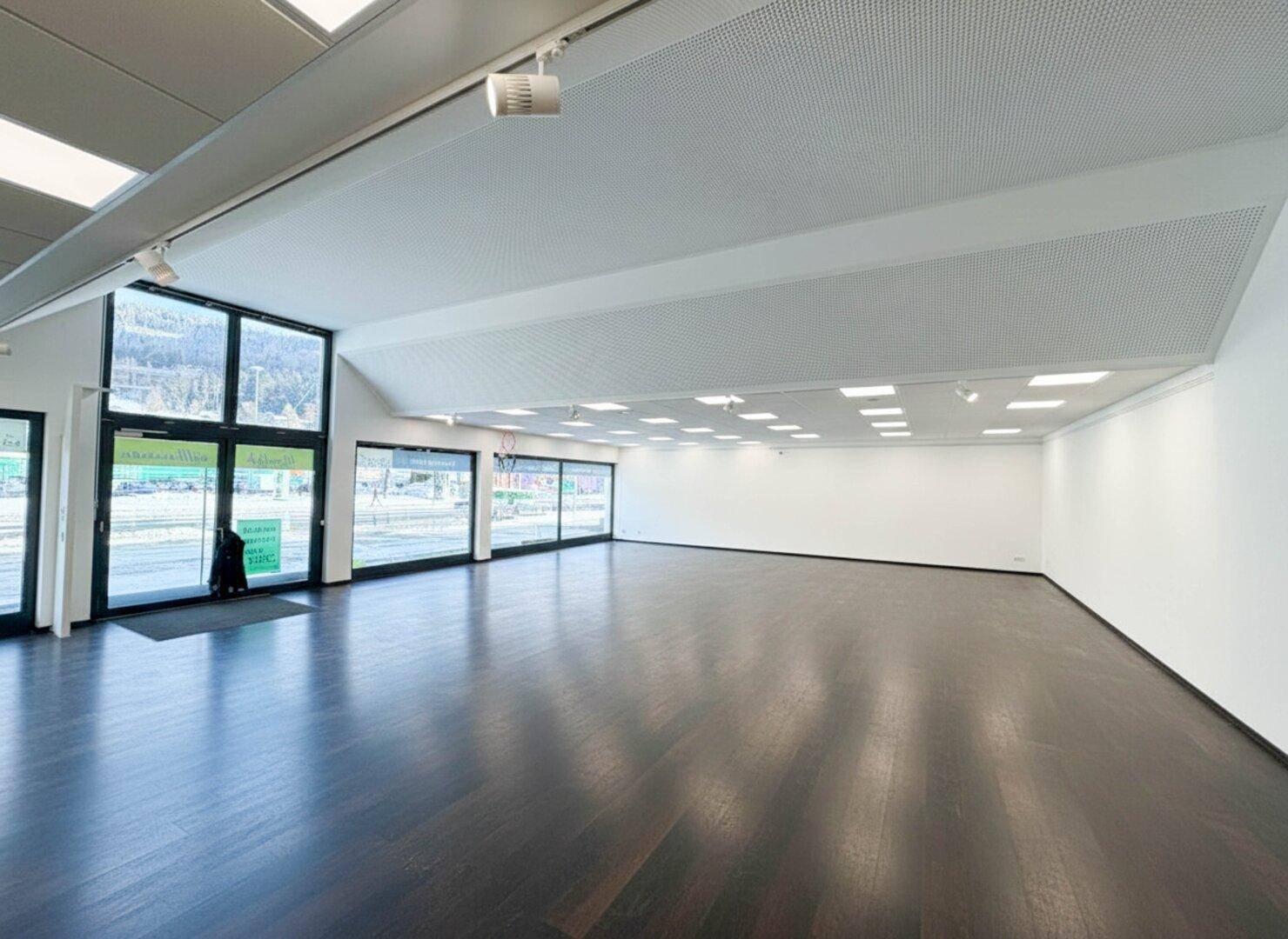 Heller Gewerberaum mit großen Fensterfronten, dunklem Boden und perforierter Akustikdecke.