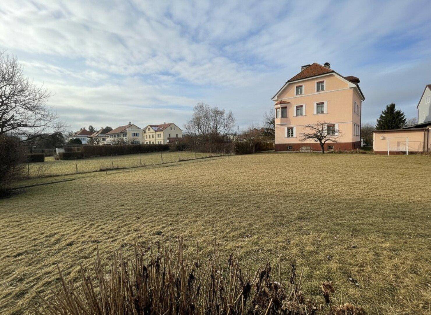 Weitläufiges Grundstück mit großem Rasenbereich und Blick auf das rosa Mehrfamilienhaus.