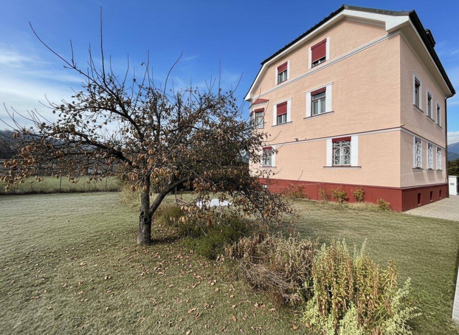 Rosa Mehrfamilienhaus mit kahlem Baum und Rasenfläche im Vordergrund unter klarem Himmel.