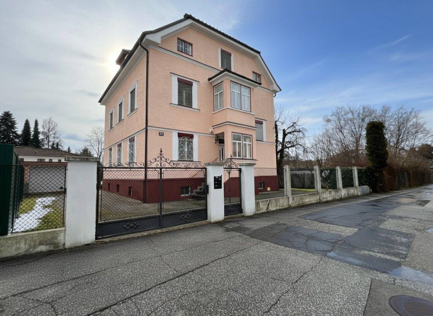 Rosa Mehrfamilienhaus mit schmiedeeisernem Tor und gepflasterter Straße im Vordergrund.
