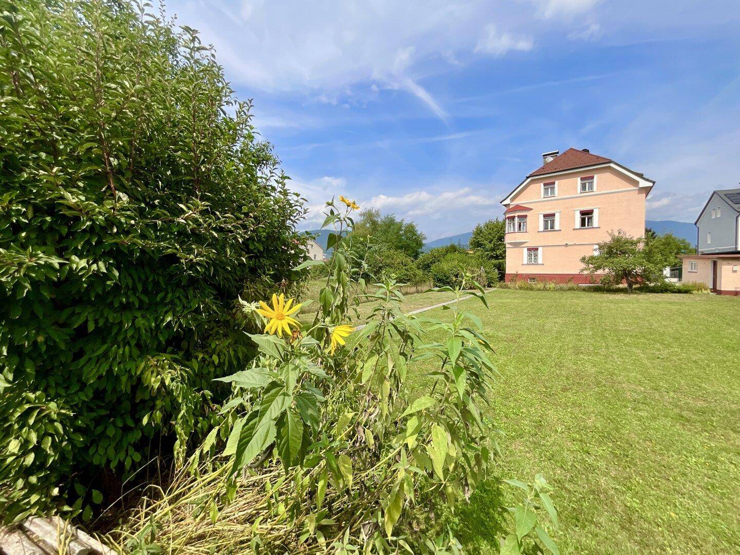 Großer Gartenbereich mit blühenden Pflanzen und dem rosa Mehrfamilienhaus im Hintergrund.