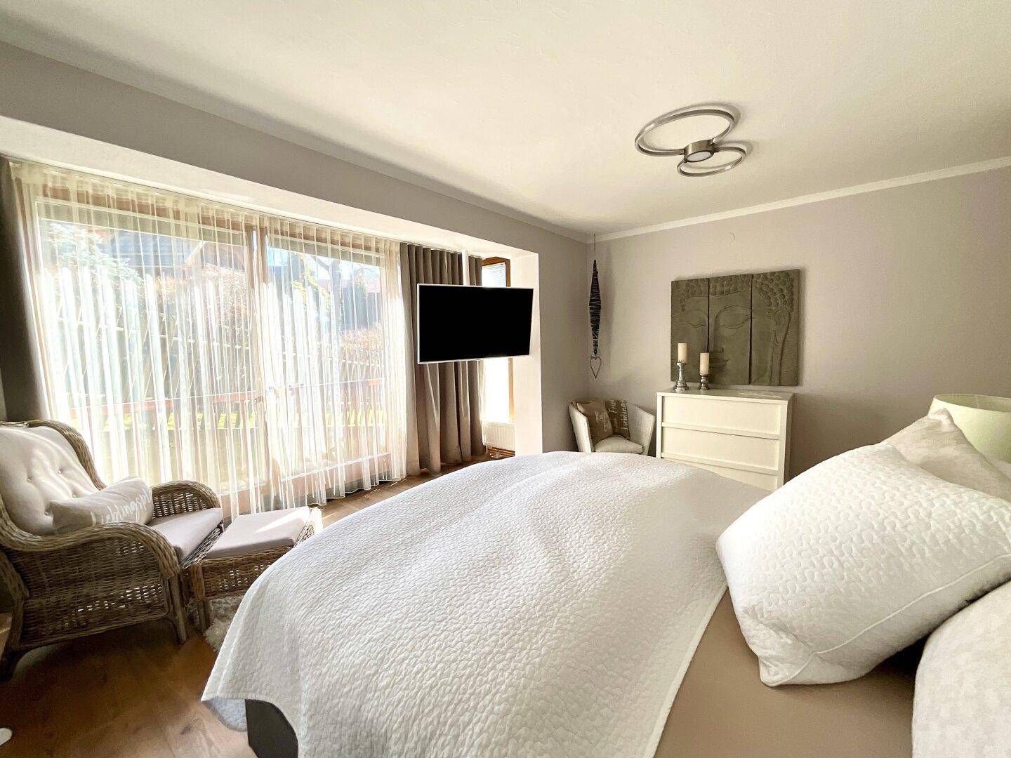 Gemütliches Schlafzimmer mit großem Fenster, hellem Bett und Rattan-Sessel.