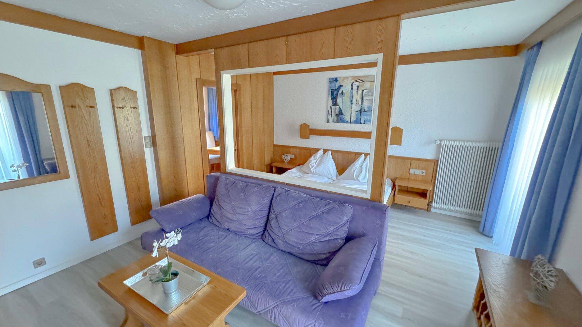 Helles Schlafzimmer mit lila Sofa, Holzelementen und separatem Schlafbereich.