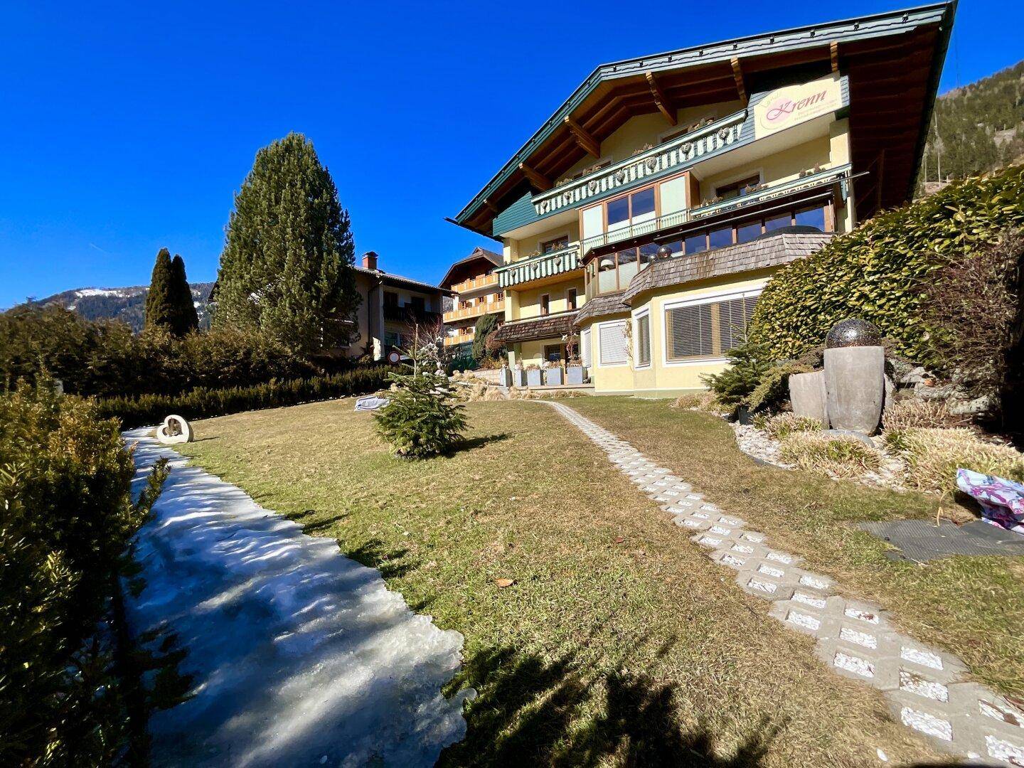 Traditionelles Gebäude mit alpiner Architektur, umgeben von einem weitläufigen Garten mit Wegen und Bäumen.