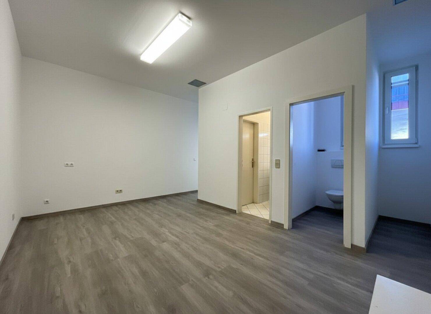 Kleinerer Raum mit Zugang zu einer Toilette, hellem Laminatboden und einem Fenster.