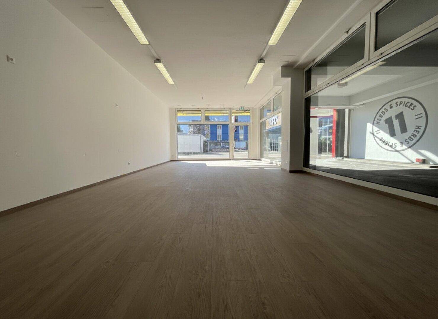 Heller Geschäftsraum mit großen Schaufenstern, Holzoptikboden und Blick auf den Eingangsbereich.