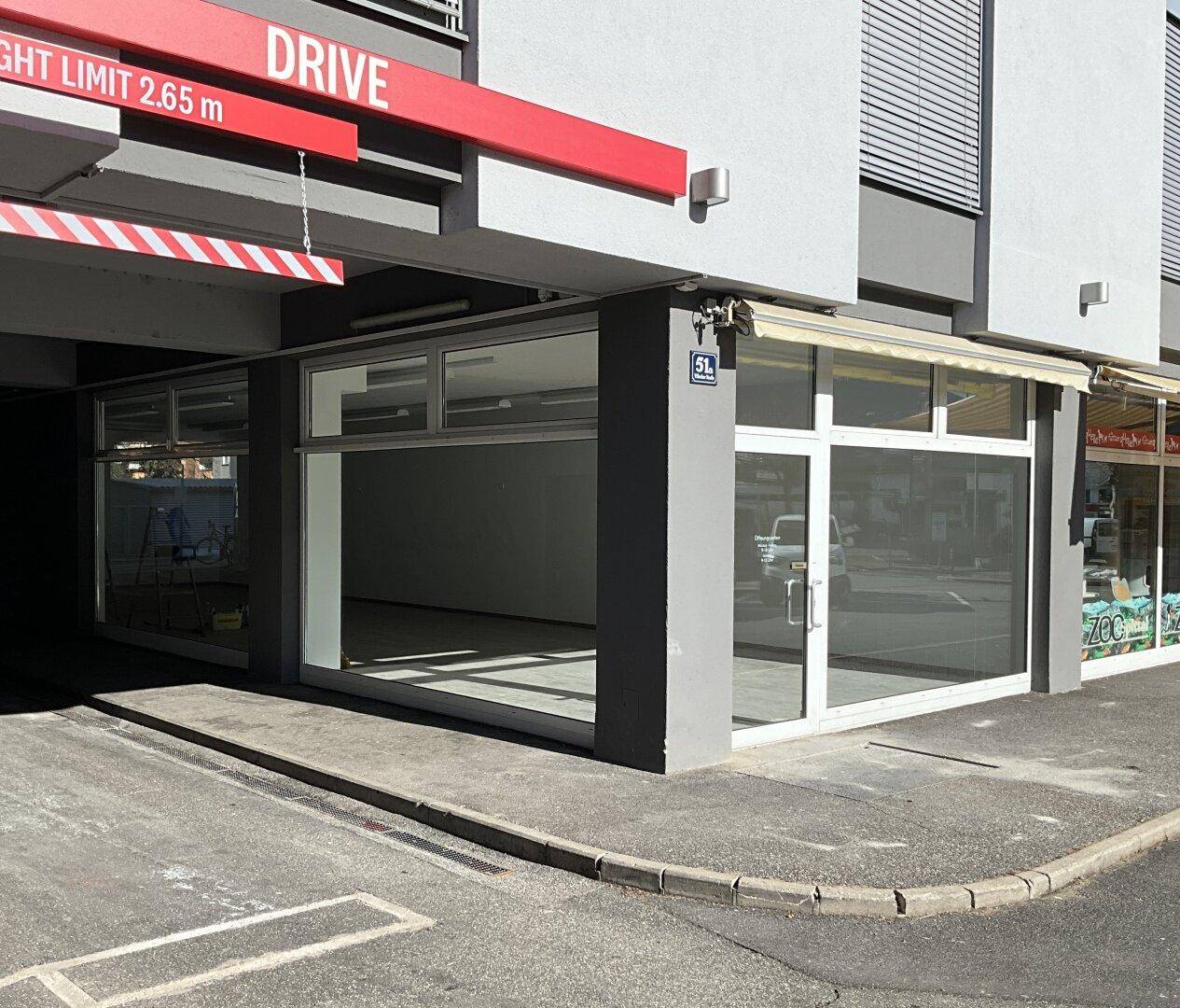 Außenansicht des Gebäudes mit Geschäftsfront und Zufahrt zu einer Tiefgarage oder einem Parkbereich.