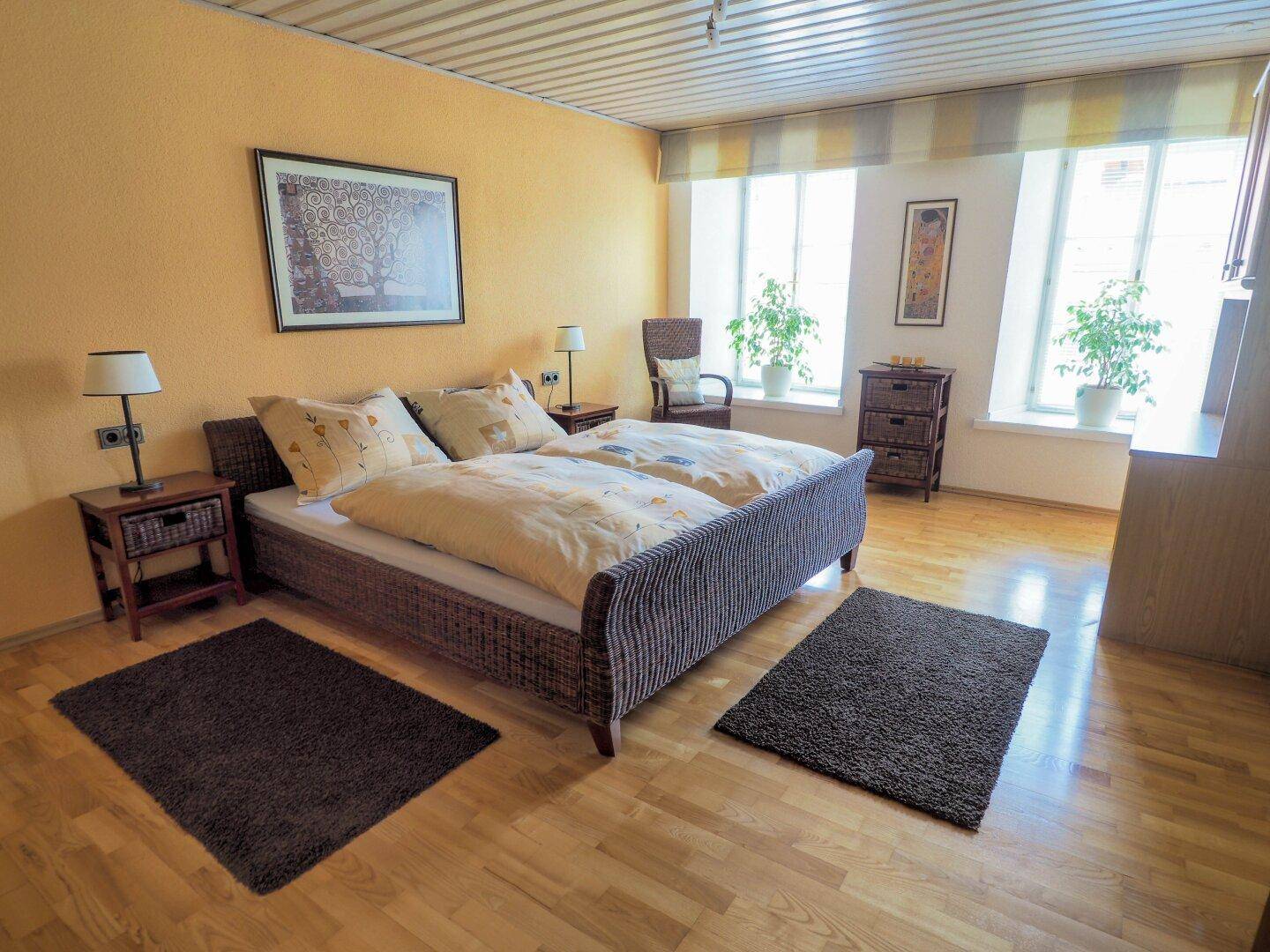 Helles Schlafzimmer mit Doppelbett, Holzboden und gelben Wänden, gut beleuchtet durch große Fenster.