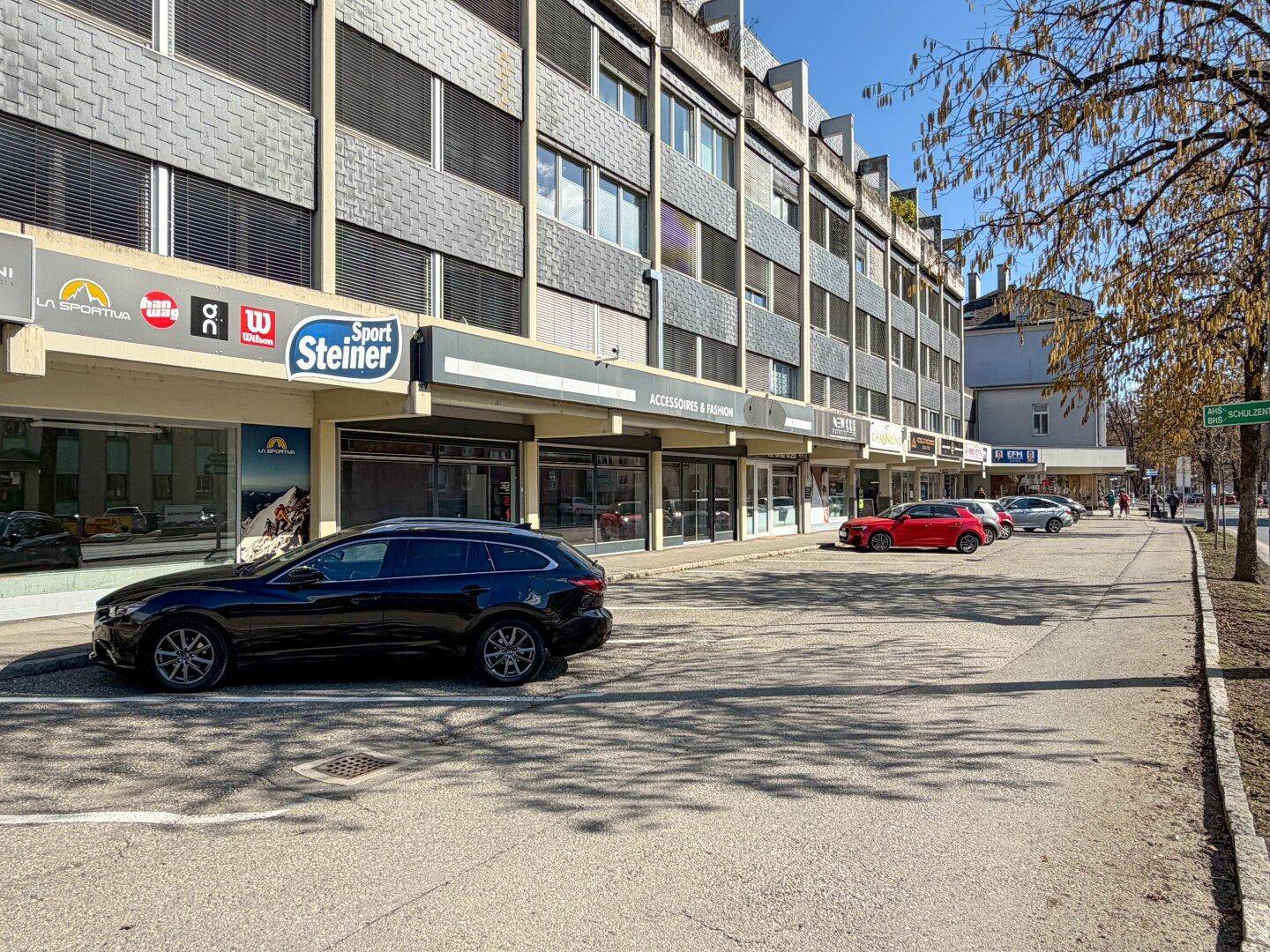Ansicht eines Geschäftsgebäudes mit verschiedenen Ladenfronten und parkenden Autos auf der Straße.