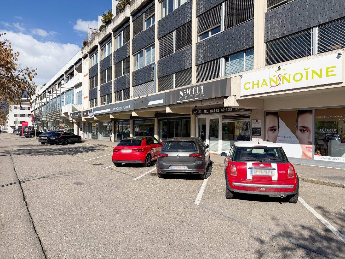 Fassade eines Geschäftsgebäudes mit mehreren Ladenfronten und parkenden Fahrzeugen davor an einem sonnigen Tag.