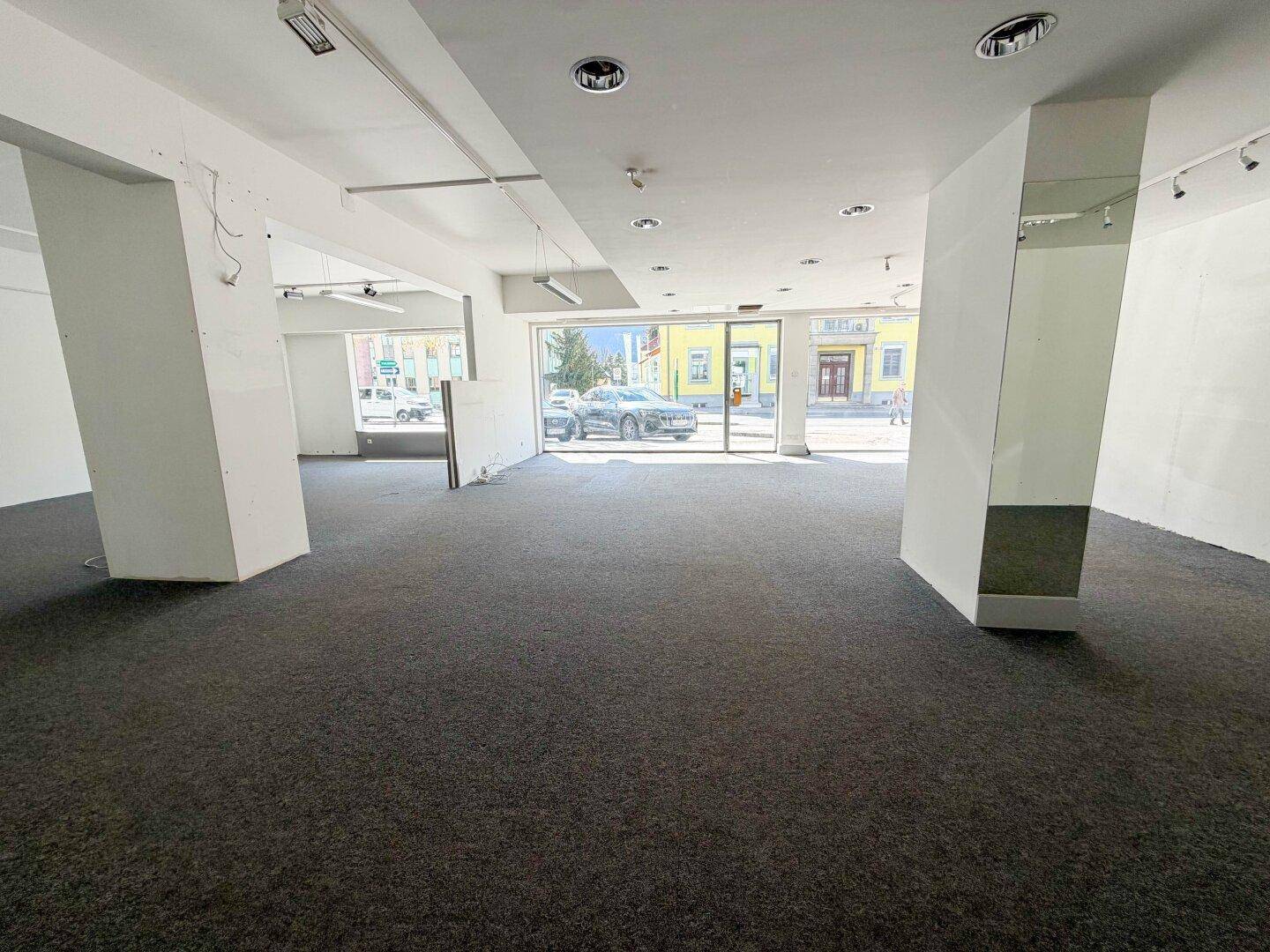 Offener Ladenbereich mit Teppichboden, weißen Wänden und großen Fenstern zur Straßenseite, sehr hell.