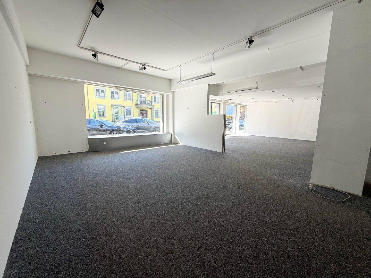 Heller Ladenraum mit Teppichboden und großen Fenstern, die viel Tageslicht hereinlassen, sehr einladend.