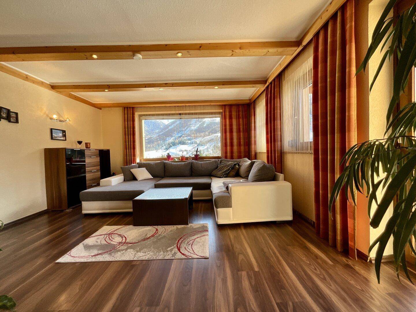 Gemütliches Wohnzimmer mit großem Ecksofa, Holzbalken und Bergblick durch das Fenster.