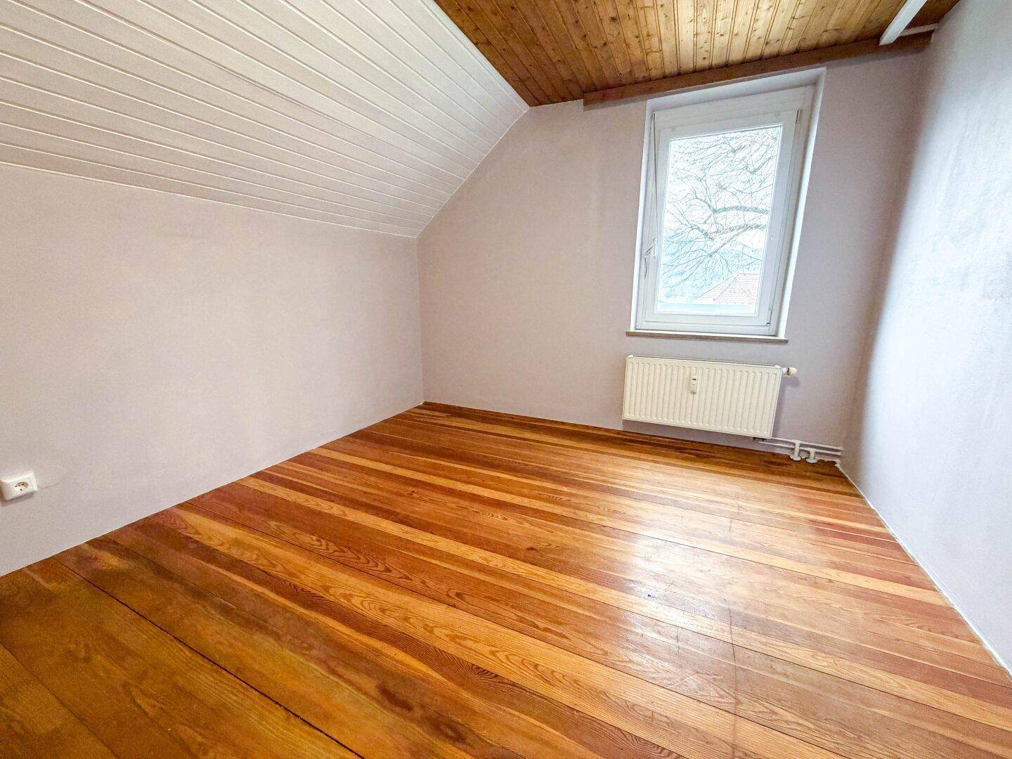 Helles Zimmer im Dachgeschoss mit Holzdecken, Holzboden und einem Fenster mit Blick ins Grüne.