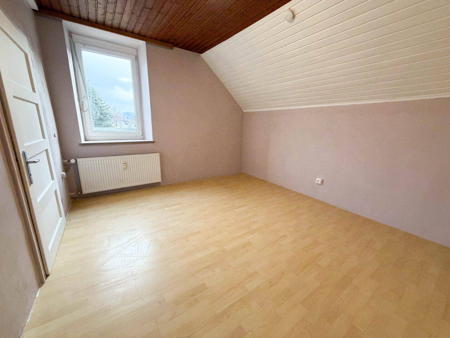 Helles Zimmer im Dachgeschoss mit Laminatboden, Holzdecken und einem Fenster.